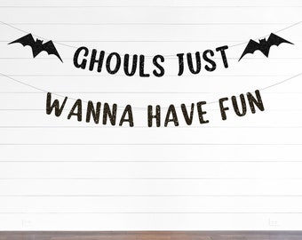May include: A black glittery banner featuring two bat silhouettes at the ends and the text GHOULS JUST WANNA HAVE FUN arranged in two lines on thin strings. The letters and bat shapes have a textured, sparkling finish against a plain background.
