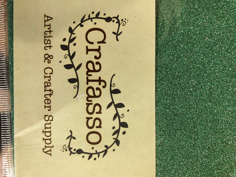 May include: A package of fine green glitter with a beige label featuring the text Crafasso Artist & Crafter Supply in dark brown font, surrounded by a decorative vine illustration. The glitter has a vibrant, reflective, and textured surface.