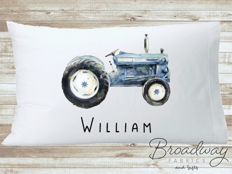 K&ouml;nnte beinhalten: Ein wei&szlig;er Kissenbezug mit einer Aquarell-Illustration eines Vintage-Traktors in Blau- und Gr&uuml;nt&ouml;nen. Der Traktor tr&auml;gt den Schriftzug "FORD" an der Seite. Der Name "WILLIAM" ist unter dem Traktor gedruckt.