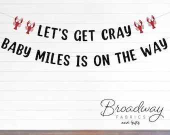 Puede incluir: Una pancarta en blanco y negro con recortes de langostas rojas dice "Let's Get Cray Baby Miles Is On The Way". La pancarta está colgada en una pared de madera blanca. El texto "Broadway Fabrics and Gifts" está en cursiva negra en la parte inferior de la imagen.