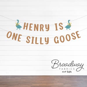 K&ouml;nnte beinhalten: Ein braunes Banner mit dem Text "Henry is one silly goose" und zwei blauen G&auml;nse-Illustrationen mit gelben Schleifen. Das Banner h&auml;ngt an einer wei&szlig;en Holztafelwand. Der Text "Broadway Fabrics and Gifts" ist unten im Bild in Schwarz gedruckt.
