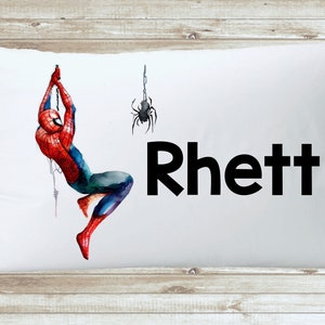 May include: White pillowcase featuring a red and blue Spiderman illustration hanging from a web. A black spider is positioned above him. The name "Rhett" is printed in black below the illustration.