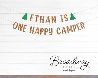 May include: A decorative banner featuring two rows of text suspended on thin strings. The top row reads ETHAN IS with a green glitter pine tree on each end. The bottom row reads ONE HAPPY CAMPER. All letters are in a brown glitter finish.