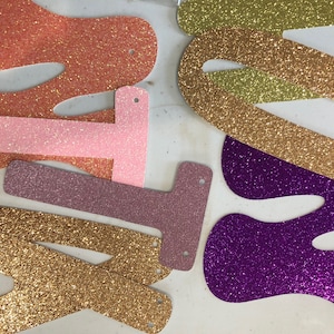 May include: A collection of glittery, die-cut letter shapes in various colors including orange, light pink, mauve, gold, and purple. Each letter features small pre-punched holes at the top and bottom, designed for stringing together to create a banner.