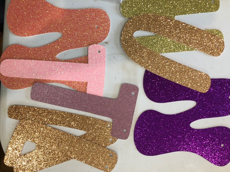 May include: A collection of glittery, die-cut letter shapes in various colors including orange, light pink, mauve, gold, and purple. Each letter features small pre-punched holes at the top and bottom, designed for stringing together to create a banner.