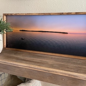 May include: A framed photograph of a sunset over a body of water with a small island in the distance. The frame is made of dark wood and the photograph is printed on canvas.