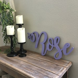 May include: A wooden sign with the word "Rose" painted in a light purple color. The sign is sitting on a rustic wooden table with three candlesticks and candles.