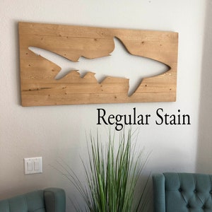 May include: A wooden wall art piece featuring a cutout of a shark silhouette. The wood is stained a light brown colour. The text "Regular Stain" is printed below the art piece.