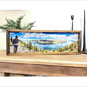 May include: A rectangular wooden frame containing a panoramic photograph of a person standing on a rocky cliff overlooking a large blue lake surrounded by evergreen trees and mountains under a bright blue sky with white clouds.