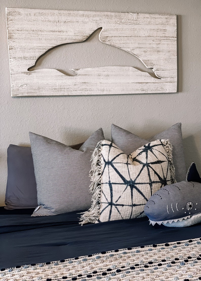 May include: A bedroom with a white wooden wall hanging featuring a cutout dolphin silhouette. The bed is made with a dark blue duvet and a patterned throw blanket. There are several pillows on the bed, including a striped pillow, a geometric patterned pillow, and a grey shark plush toy.