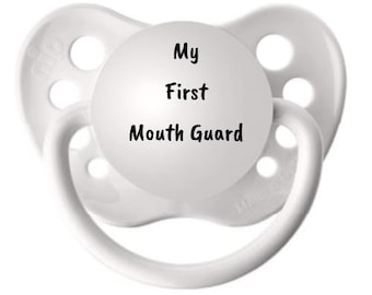 May include: A white pacifier featuring a circular center plate with the black printed text My First Mouth Guard. The pacifier has a shield with four ventilation holes and a curved handle attached to the front, all in a solid white finish.