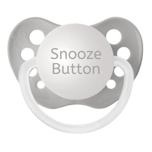 May include: A light gray pacifier featuring a circular white center with the text Snooze Button printed in gray. The shield has six ventilation holes and a translucent white ring handle attached to the front for easy gripping and handling.