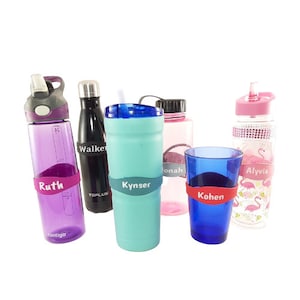 May include: Six personalized water bottles with different names on them. The bottles are in various colors, including purple, black, turquoise, pink, and blue. The bottles are all made of plastic and have a variety of designs.