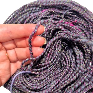 Puede incluir: Un ovillo de hilo en tonos de morado, gris y rosa con hilo metálico dorado tejido en todo.