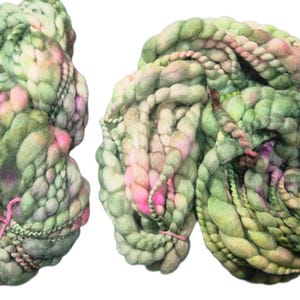 Puede incluir: Dos madejas de hilo teñido a mano en tonos verdes, rosas y crema. El hilo tiene un aspecto grueso y texturizado, con una construcción trenzada y gruesa. Un hilo rosa asegura cada madeja. Ideal para proyectos de tejido o ganchillo.