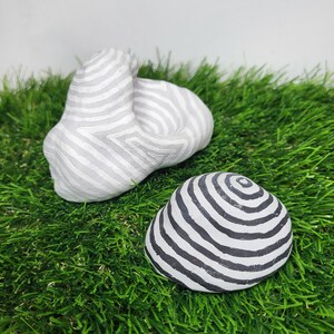 Pode incluir: Duas pedras decorativas de cerâmica com padrões de listras pretas e brancas. A pedra maior tem uma forma mais abstrata, enquanto a pedra menor é redonda. Ambas as pedras estão sobre um leito de grama artificial verde.