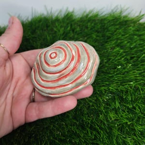 Pode incluir: Um pequeno objeto de cerâmica pintado à mão em forma de espiral com listras vermelhas e brancas sobre um fundo cinza.