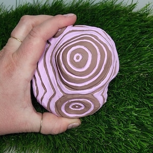 Pode incluir: Uma mão segura uma escultura de cerâmica rosa e marrom com um design de círculos concêntricos.