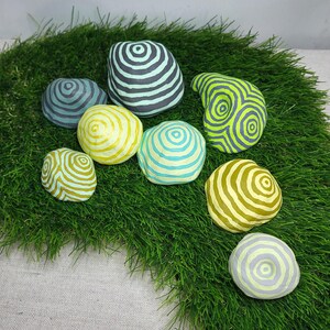 Pode incluir: Um conjunto de oito pedras decorativas pintadas com círculos concêntricos em tons de verde, amarelo e azul. As pedras estão dispostas sobre um leito de grama artificial verde.