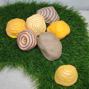 Pode incluir: Sete pedras pintadas com diferentes padrões de círculos concêntricos amarelos, rosa, laranja, azuis e cinzentos. As pedras estão dispostas sobre um leito de grama artificial verde.