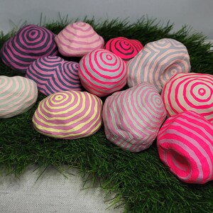Pode incluir: Uma coleção de 10 pedras decorativas redondas pintadas à mão com círculos concêntricos rosa, amarelos e cinzentos. As pedras estão dispostas sobre um leito de grama artificial verde.