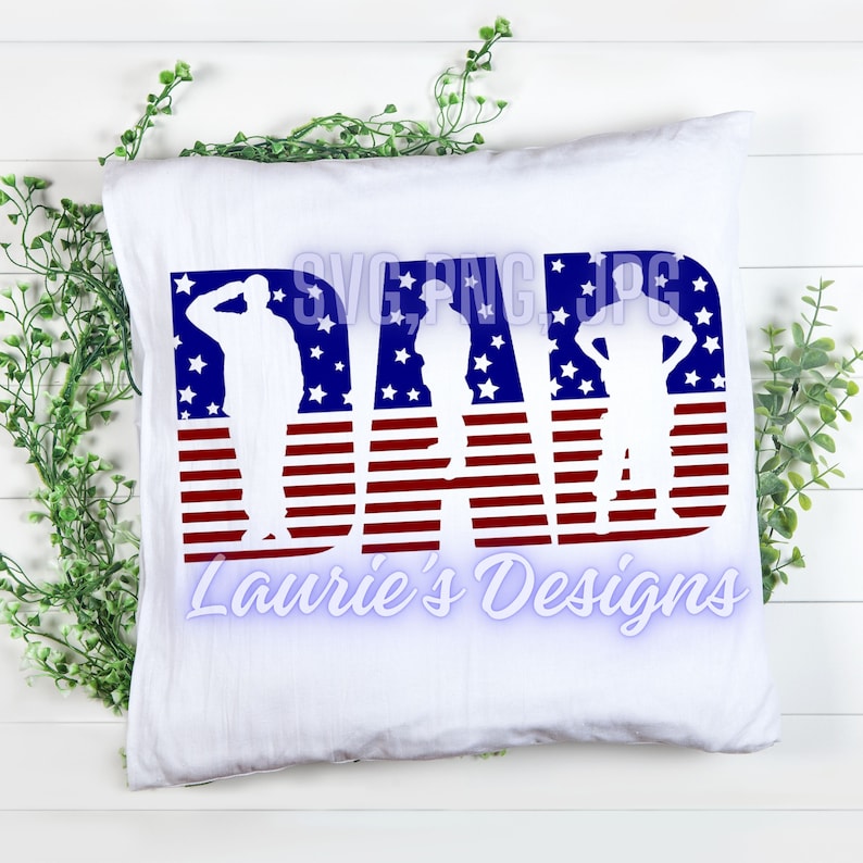 May include: A white pillow with the word "DAD" in red, white, and blue. The letters are made up of American flag stripes and stars. There are three silhouette figures of soldiers inside the letters. The text "Laurie's Designs" is below the word "DAD".