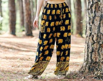 Puede incluir: Una mujer con un top blanco corto y pantalones negros con estampado de elefantes dorados se encuentra en un bosque de pinos. Se apoya en un gran pino.