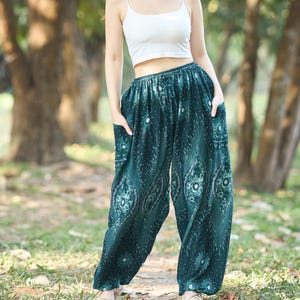 May include: A woman wearing a white crop top and teal patterned pants with pockets. The pants have a decorative design. The background is a park with trees and grass.
