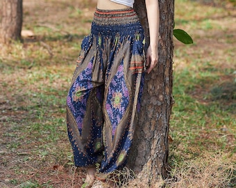 May include: A woman wearing a white crop top and blue and gold patterned pants stands in a forest with her arms raised above her head. She is leaning against a tree trunk.