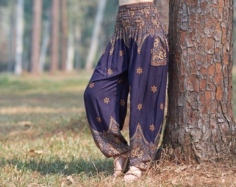 May include: A woman wearing a white crop top and blue printed pants stands in a forest, leaning against a tree.