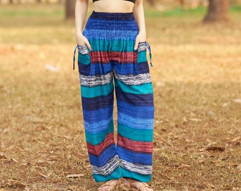 May include: A woman wearing a black crop top and blue, red, and teal striped pants. The pants have a drawstring waist and pockets. The background is a forest with tall trees and brown leaves on the ground.