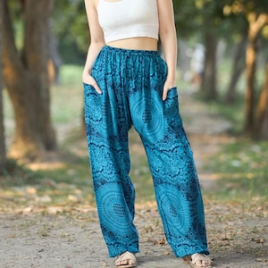 Puede incluir: Una persona lleva un top corto blanco y pantalones azules estampados con bolsillos. Los pantalones tienen un diseño tipo mandala. El fondo es una escena exterior borrosa con árboles.