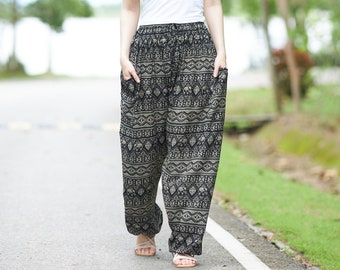 May include: A woman wearing a white tank top and black and beige patterned pants with a geometric design. She is walking on a paved path with green trees in the background.