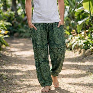 May include: A person wearing a white t-shirt and green patterned pants walks on a path surrounded by lush greenery. The pants have a dark green paisley design. The person is barefoot.