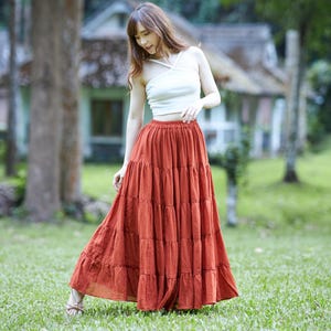 Może przedstawiać: Biały bezrękawnik typu crop top ze skrzyżowanym dekoltem w połączeniu z długą, warstwową sp&oacute;dnicą maxi w kolorze rdzawego pomarańczu. Sp&oacute;dnica posiada marszczony elastyczny pas oraz pełną, zwiewną sylwetkę sięgającą kostek, wykonaną z tkaniny.