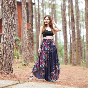 May include: A woman wearing a black crop top and a long, flowing floral skirt stands in a pine forest. The skirt has a dark blue base with pink, purple, and green floral patterns. The woman is looking to the right of the frame.