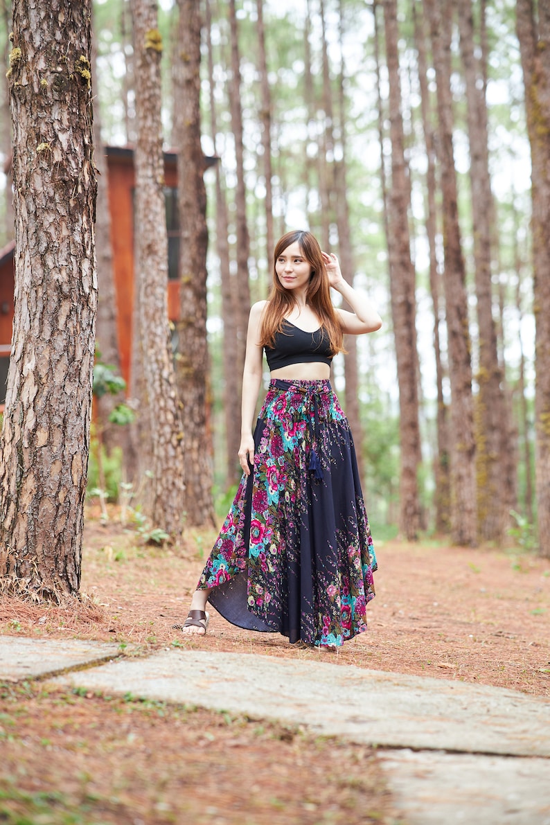 May include: A woman wearing a black crop top and a long, flowing floral skirt stands in a pine forest. The skirt has a dark blue base with pink, purple, and green floral patterns. The woman is looking to the right of the frame.
