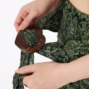 Op de afbeelding: Een donkergroen kledingstuk van stof met een herhalend zwart bloemen- en geometrisch patroon, vastgezet in de taille met een grote, hartvormige, donkerbruine, getextureerde houten gesp waar de stof doorheen is gehaald.