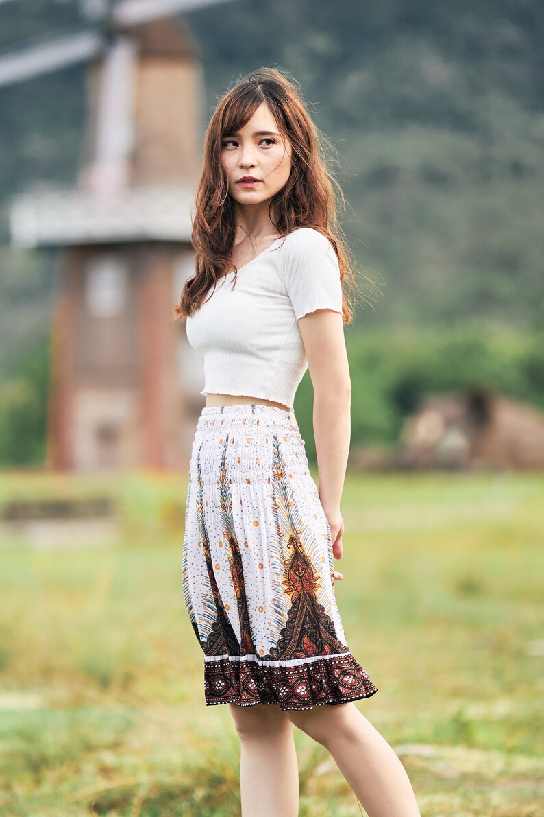 K&ouml;nnte beinhalten: Eine Frau tr&auml;gt ein wei&szlig;es Crop-Top und einen wei&szlig;en Rock mit braunem Muster. Sie steht auf einem Feld mit gr&uuml;nem Gras.