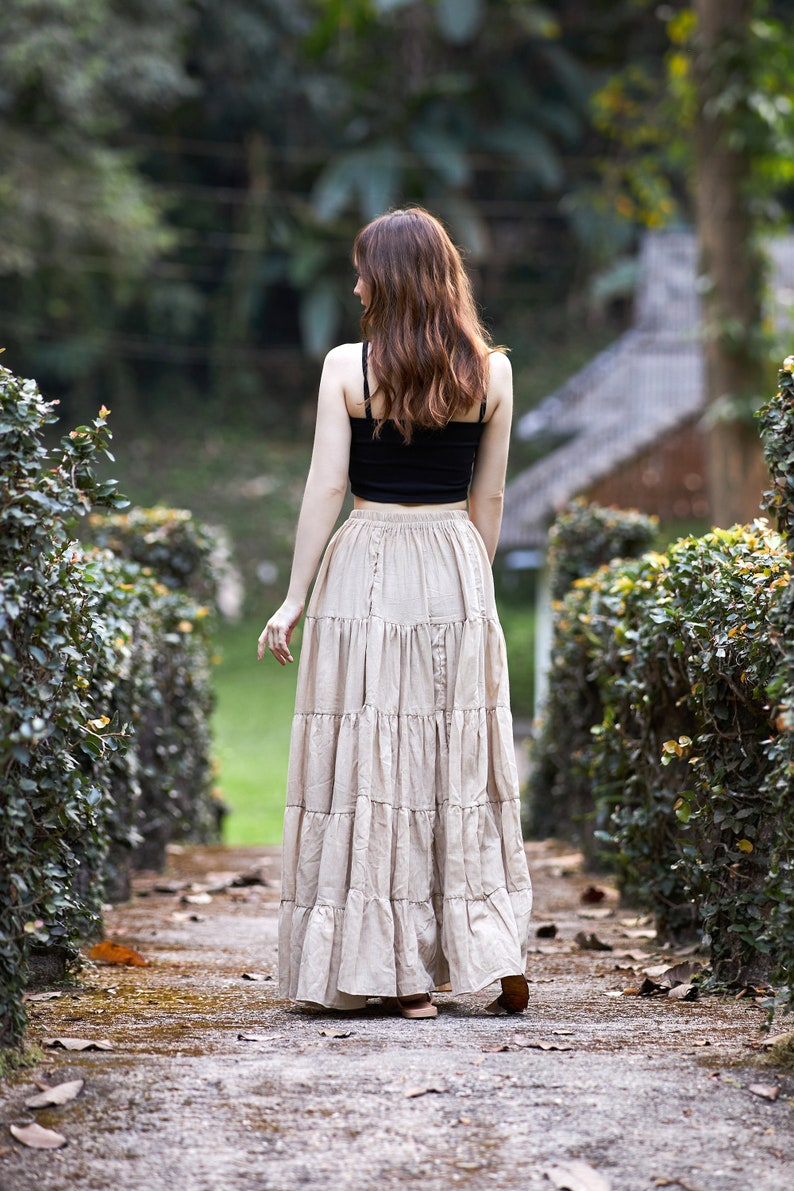 May include: A woman wearing a black tank top and a long, tiered beige skirt walks down a path lined with green bushes.