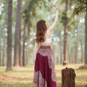 Puede incluir: Una mujer con un top blanco corto y una falda larga de color burdeos con un patr&oacute;n de mandala blanco se encuentra en un bosque, alcanzando una rama de &aacute;rbol.