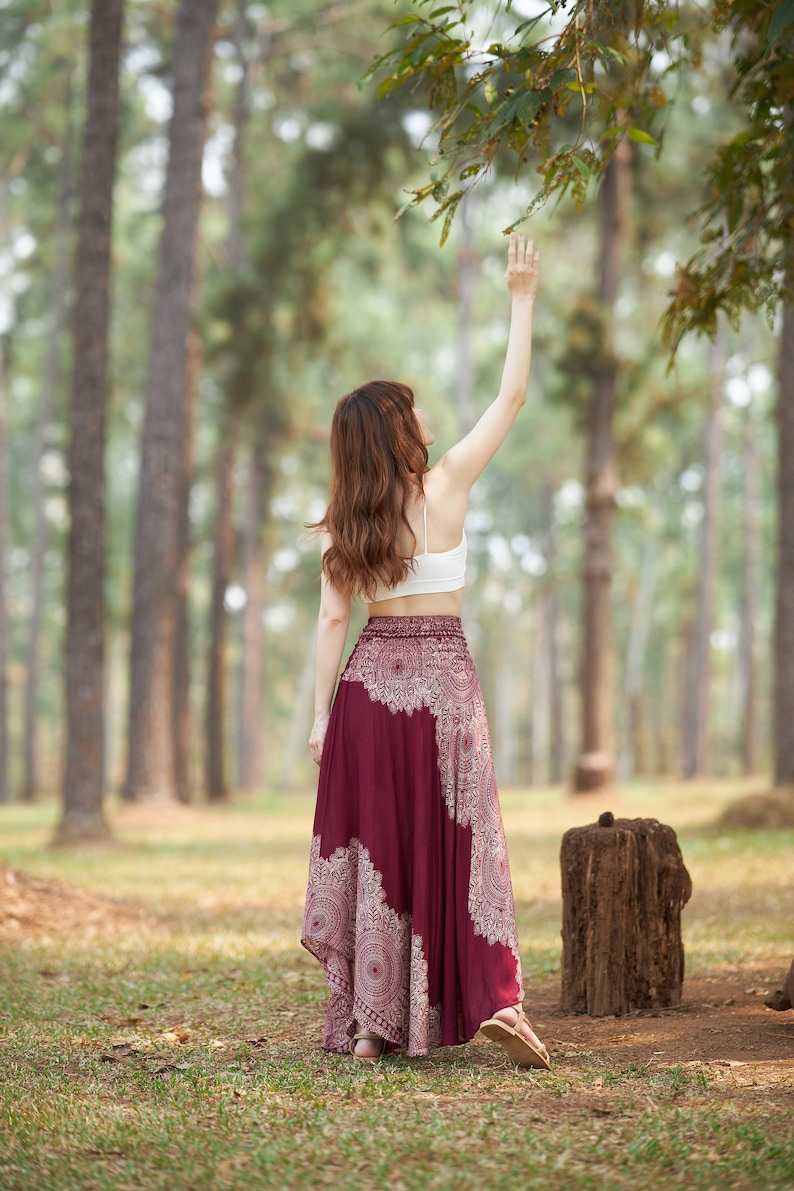 Puede incluir: Una mujer con un top blanco corto y una falda larga de color burdeos con un patr&oacute;n de mandala blanco se encuentra en un bosque, alcanzando una rama de &aacute;rbol.