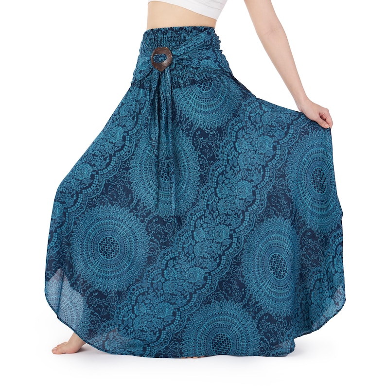 Op de afbeelding: Een enkellange rok in blauwgroen en donkerblauw met een herhalend cirkelvormig mandalapatroon en diagonale banen met bloemachtige kantmotieven. De rok heeft een hoge, gesmokte elastische tailleband met een centraal donkerbruin rond accentstuk.