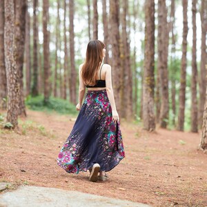 May include: A woman wearing a black tank top and a long, flowing floral skirt walks through a pine forest.