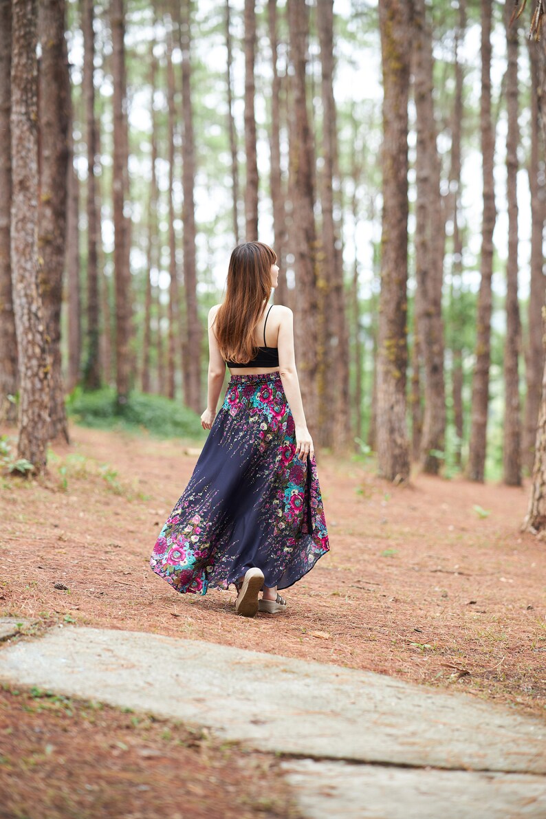 May include: A woman wearing a black tank top and a long, flowing floral skirt walks through a pine forest.
