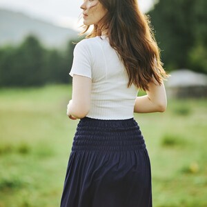 Op de afbeelding: Een vrouw draagt een wit shirt met korte mouwen en een donkerblauwe rok met een geruite zoom. Ze staat in een veld met groen gras.