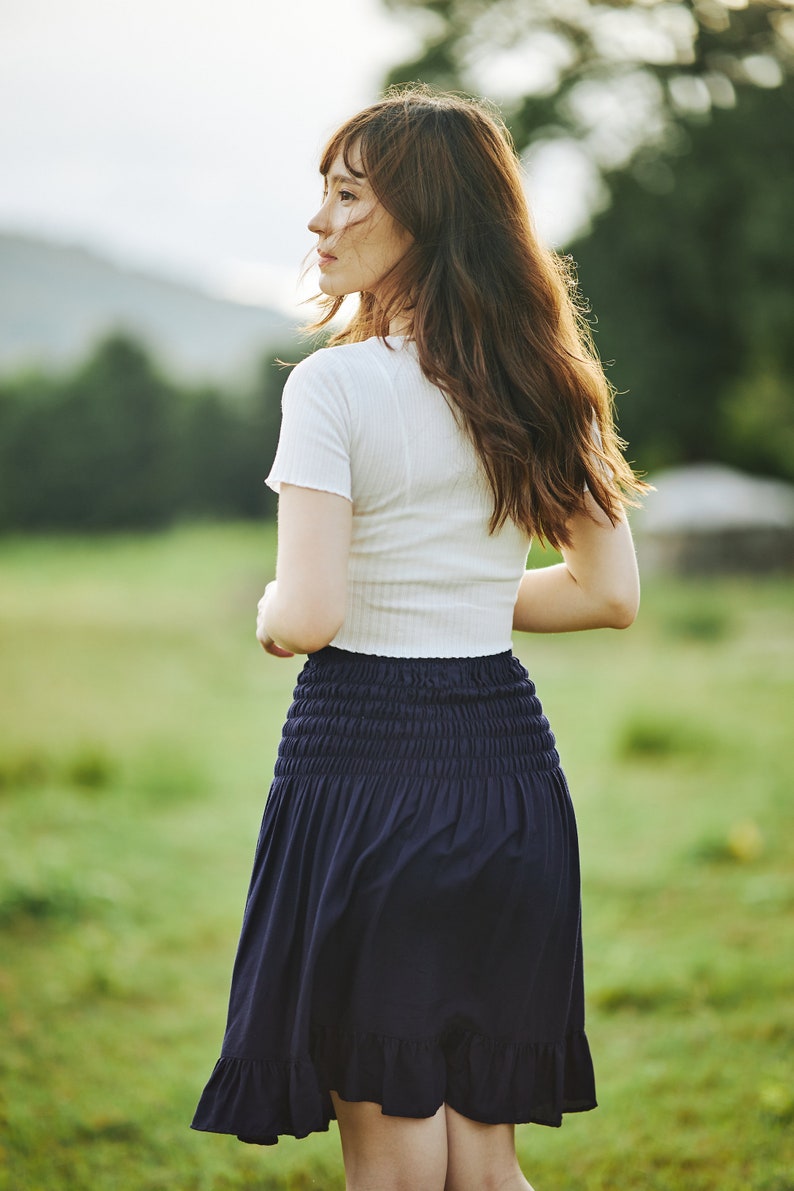Op de afbeelding: Een vrouw draagt een wit shirt met korte mouwen en een donkerblauwe rok met een geruite zoom. Ze staat in een veld met groen gras.