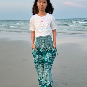 May include: A young person wearing a white t-shirt with the text "HAPPY FUNNY" printed on it, and green pants with an elephant print. The person is standing on a sandy beach with the ocean in the background.