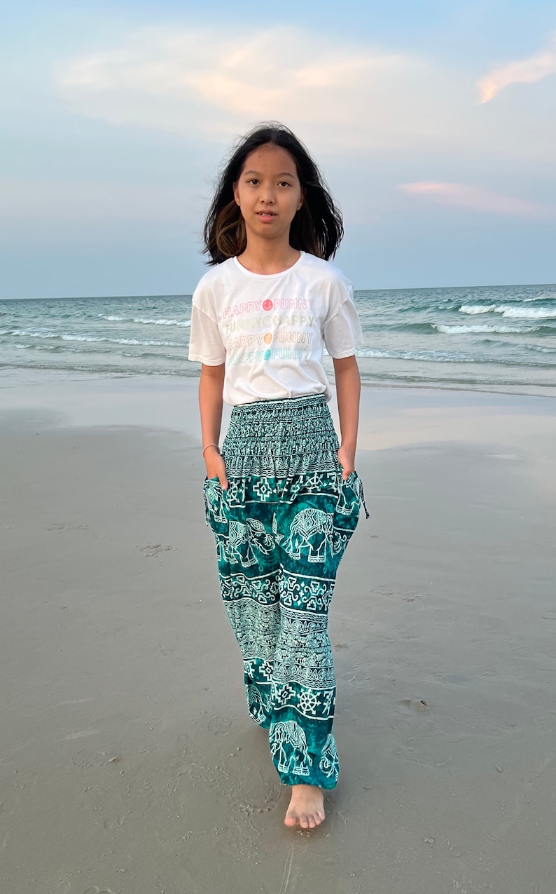 May include: A young person wearing a white t-shirt with the text "HAPPY FUNNY" printed on it, and green pants with an elephant print. The person is standing on a sandy beach with the ocean in the background.