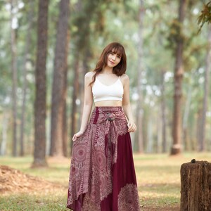 Puede incluir: Una mujer con un top blanco corto y una falda larga de color burdeos con un dise&ntilde;o estampado. La falda tiene un cintur&oacute;n con una hebilla decorativa. Est&aacute; de pie en un entorno boscoso.